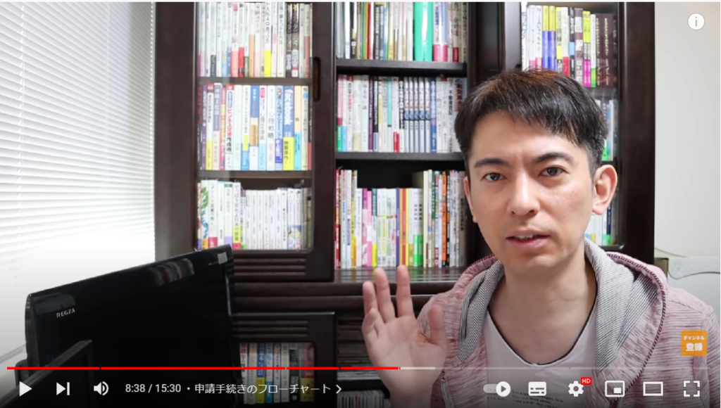 山田さんが申請方法のフローチャートについて解説し始める場面。山田さんは、本棚の前に座っている。
