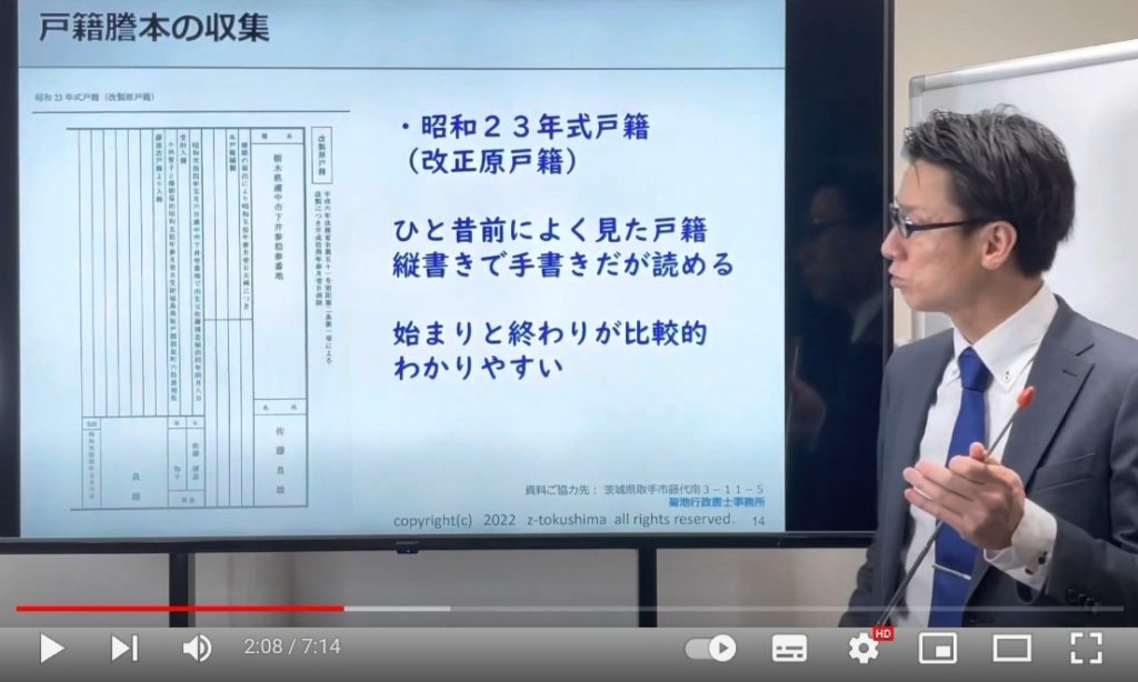 会議室のようなところが映っていて、画面右側には尾崎さんがいらっしゃいます。
画面左側には、｢昭和23年式戸籍｣の見本とその解説が映された大きなスクリーンが設置されています。
(このスクリーンはとても大きく画面の3分の2くらいを占めています)

この場面で尾崎さんは、｢昭和23年式戸籍｣の解説をされています。