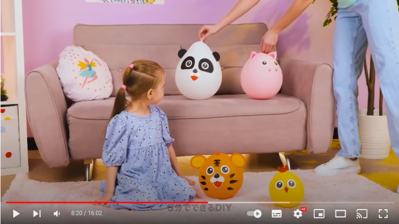 ソファーがある部屋で、ソファーの前に女の子が座っています。女の子の横には虎と鶏の風船、ソファーにはパンダと豚の風船が置いてあります。パンダと豚の風船は大人の人が置こうとしているところです。